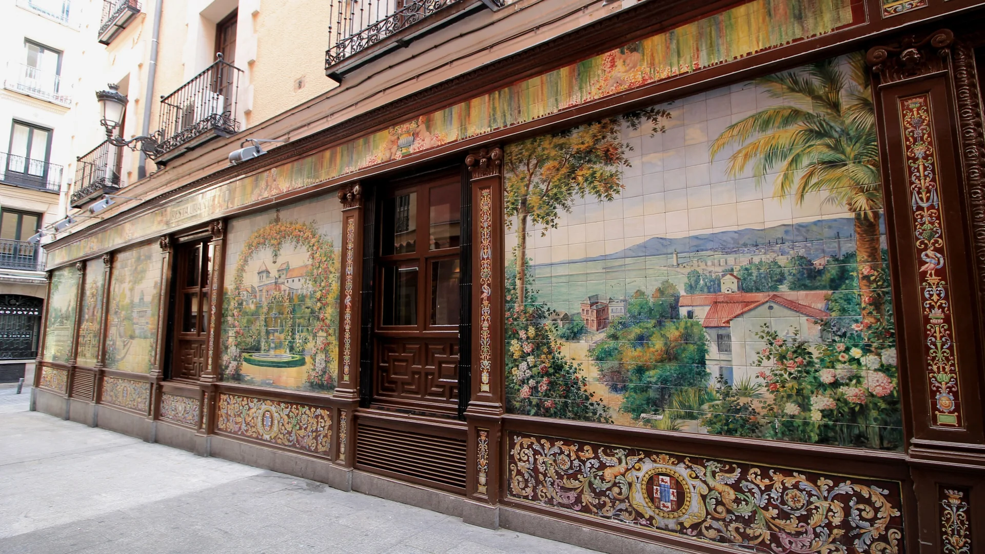 The tiled front of the former Villa Rosa on Plaza de Santa Ana in Madrid: a large painted ceramic panel of a bay with palm trees and red-roofed houses, framed in dark carved wood above a decorated tile base